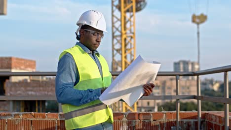 Portrait-shot-of-the-African-American-man-constructor-in-the-hardhat-and-goggles-holding-plans-and-drafts-and-studying-them-while-standing-outdoors-at-the-building-site.
